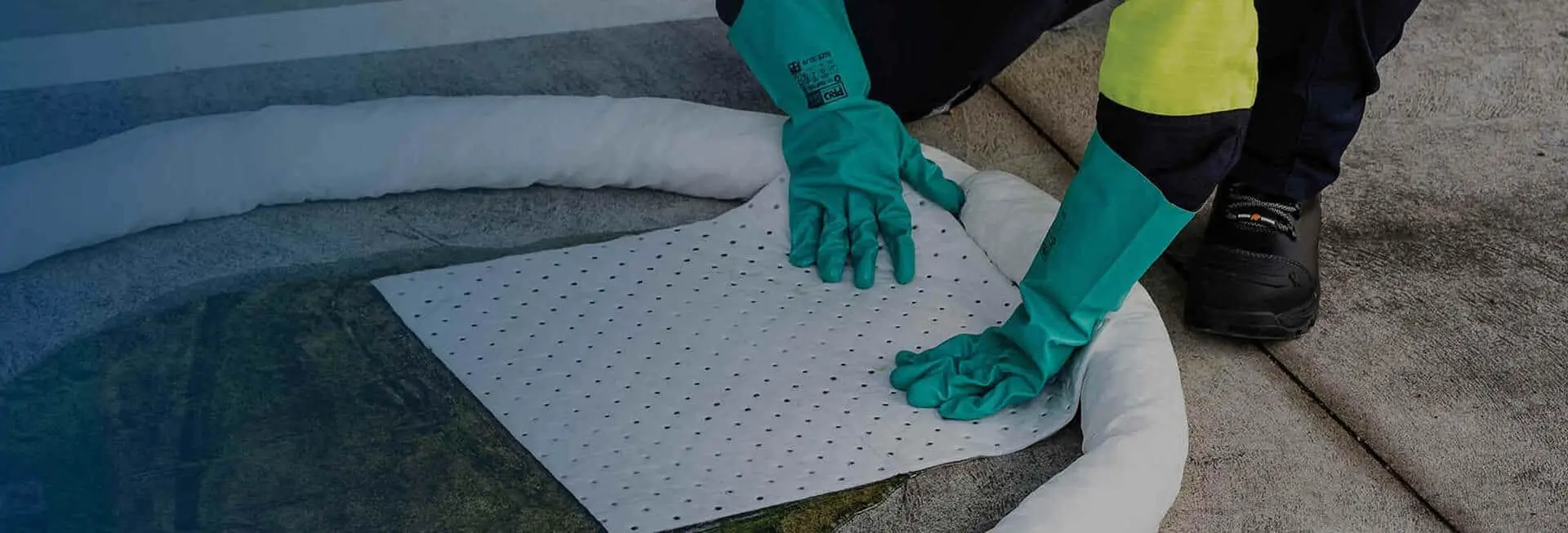 Close-up of a person in teal gloves placing a patterned absorbent mat on the ground, surrounded by materials on a work surface, indicating a safety or cleanup procedure.